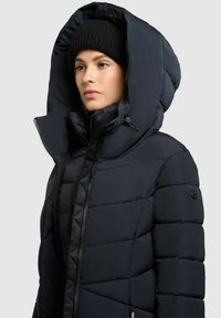 Tumši zila pūkaina jaka ar lielu kapuci, quilted tekstūru un pilnu rāvējslēdzēju. Ir uzloga akcents uz piedurknes.