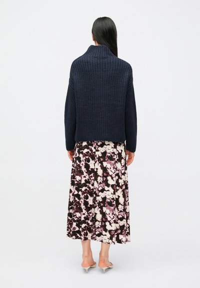 Σκούπο μπλε πλεκτό πουλόβερ με ψηλό γιακά, συνδυασμένο με μακριά φλοράλ φούστα με μωβ και κρεμ αποχρώσεις. Λείο ύφασμα, άνετη εφαρμογή.