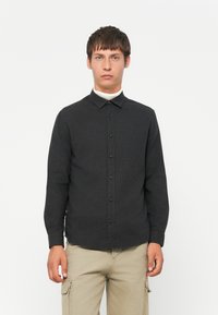 Giovane uomo che indossa una camicia a bottoni scura sopra un dolcevita bianco e pantaloni cargo beige, in piedi contro uno sfondo bianco semplice.