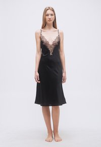 Γυναίκα που φοράει μαύρο σατέν φόρεμα με δαντελένιες λεπτομέρειες στο βαθύ V ντεκολτέ, όρθια ξυπόλητη σε απλό λευκό φόντο.