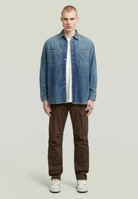 Ung man som står rak med en blå denimjacka, vit skjorta, bruna cargobyxor och vita sneakers mot en enfärgad bakgrund.