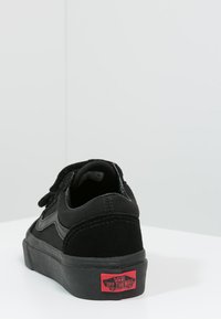 Black suede sneaker with a low profile, Velcro straps, and a textured surface. Features a red Vans logo on the back tab.