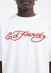 White cotton t-shirt featuring a large red rhinestone-embellished script logo reading "Ed Hardy," with short sleeves and a round neckline.