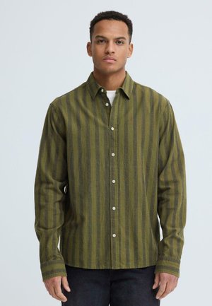 Ung mand iført en olivengrøn og mørkstribet langærmet skjorte med knapper over en hvid undertrøje, stående foran en ensfarvet baggrund.