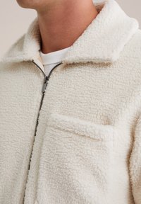 Jachetă din fleece cu fermoar, de culoare crem, cu guler, exterior texturat și un buzunar pe piept. Include accesorii din metal negru în contrast.