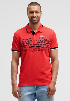 Smiling man wearing a red polo shirt with black text and light blue jeans, standing against a plain light gray background.