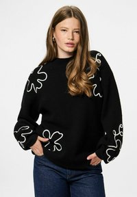 Pull en maille noire avec motifs floraux abstraits blancs sur les manches, col rond côtelé et coupe ample. Associé à un jean bleu.