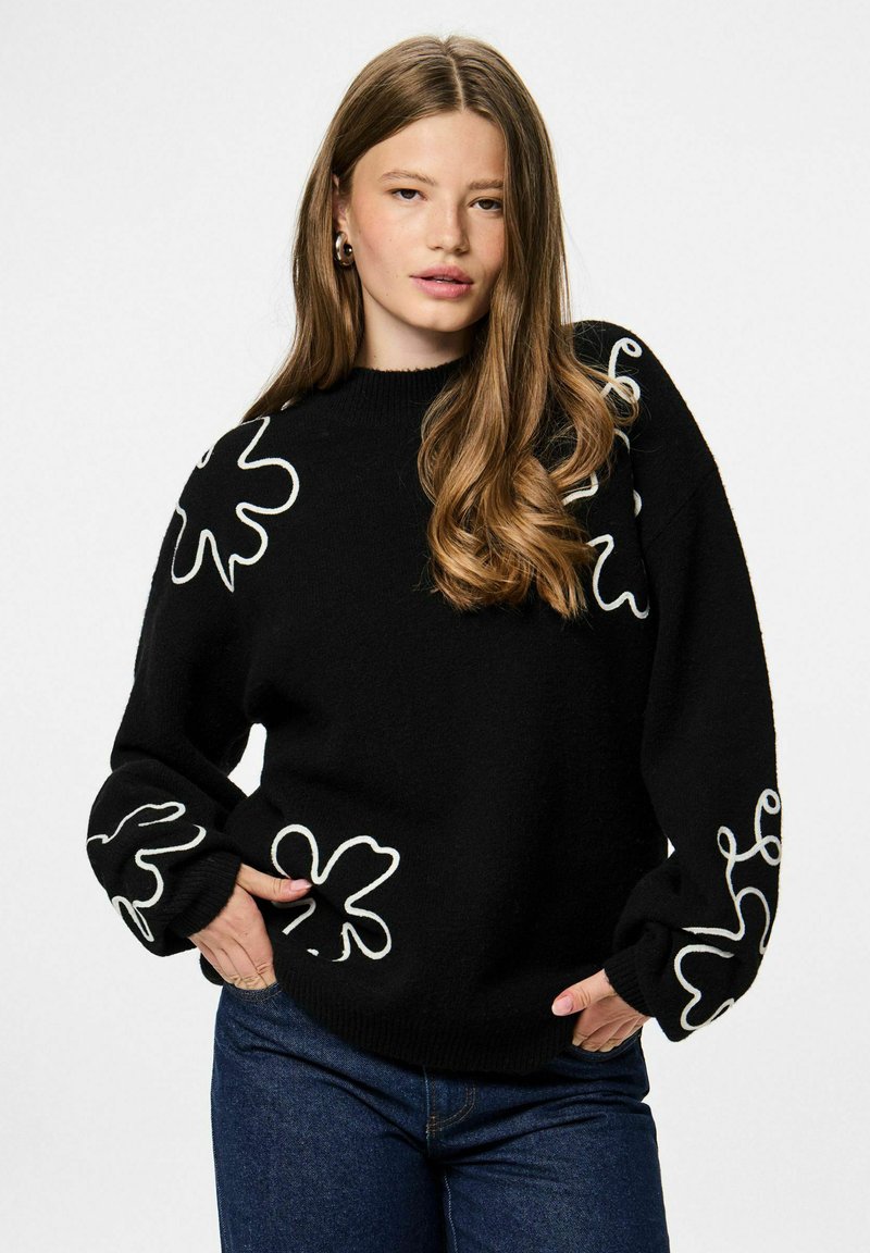 Pull en maille noire avec motifs floraux abstraits blancs sur les manches, col rond côtelé et coupe ample. Associé à un jean bleu.