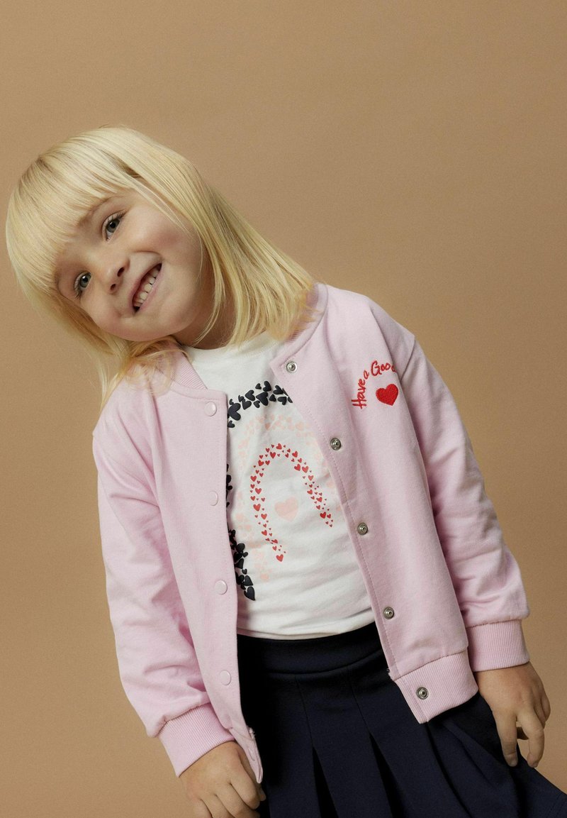 Cardigan rosa chiaro con bottoni a pressione, con la scritta "Have a Good" e un cuore rosso. Sotto, una t-shirt bianca con un design di arcobaleno a forma di cuore.