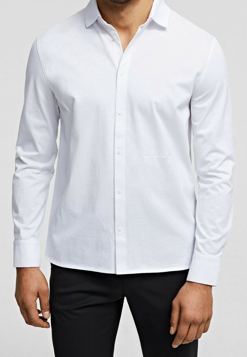 Homme portant une chemise blanche cintrée à manches longues boutonnée et un pantalon noir, debout les bras détendus le long du corps.