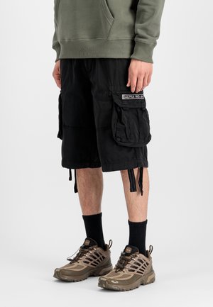 Person wearing black cargo shorts with a pocket patch, black socks, brown athletic shoes, and an olive green sweatshirt with hands relaxed.