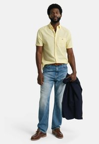 Kurzärmliges gelbes Hemd mit Knöpfen, hellblaue Jeans, braune Schuhe, hält ein marineblaues Blazer; lässige Passform mit einer Detailtasche auf der Vorderseite.