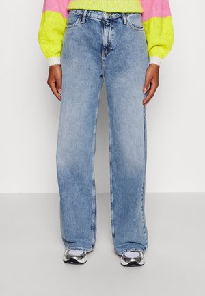 Person iført lyseblå vidde jeans, neongul og pink sweater og hvide sneakers, der står mod en ensfarvet baggrund.