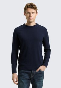 Marineblauwe trui met lange mouwen, een ronde hals, gestructureerde stof en eenvoudig ontwerp. Gecombineerd met een blauwe spijkerbroek.