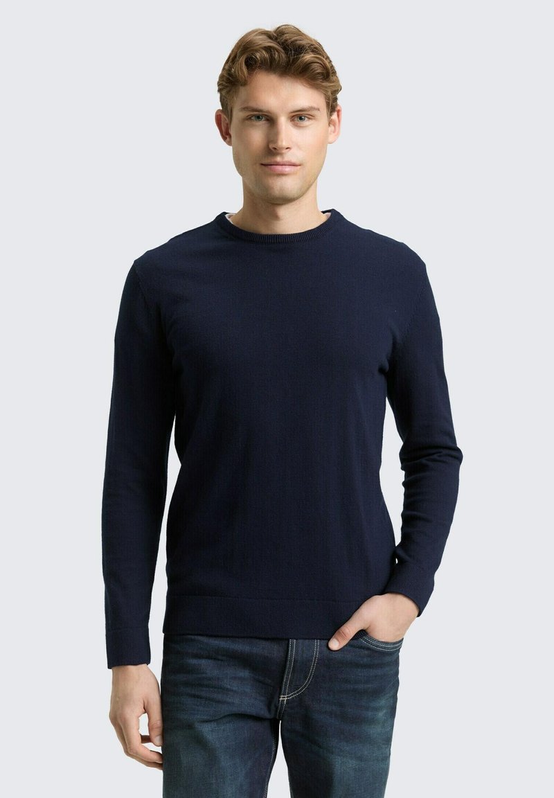 Marineblauwe trui met lange mouwen, een ronde hals, gestructureerde stof en eenvoudig ontwerp. Gecombineerd met een blauwe spijkerbroek.