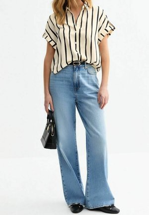 Kvinde iført en kortærmet, cremefarvet bluse med sorte lodrette striber, lyseblå vidde jeans med høj talje, sorte sko og holder en sort håndtaske.