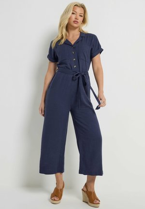 Kvinde med blondt hår iført en marineblå kortærmet jumpsuit med knapper og bælte samt beige kilehøjhælede sandaler, stående mod en hvid baggrund.