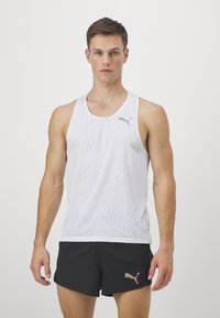 Canottiera atletica bianca senza maniche con un motivo a onde, con logo Puma in rosa. Abbinata a pantaloni corti neri, texture liscia, design sportivo.