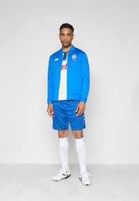 Mand iført blå sportsjakke, matchende shorts, hvide fodboldstrømper med skinnebeskyttere, hvide fodboldstøvler og en hvid trøje, stående foran en ensfarvet baggrund.