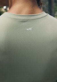 Olívazöld sportpóló kerek nyakkivágással, a felső háton kis fehér "self." logóval. Puha, sima anyagú textúra.