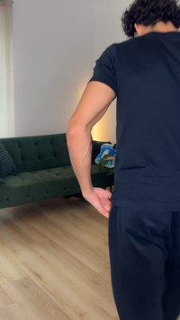 Άνδρας ντυμένος με μαύρα ρούχα στέκεται σε σαλόνι με πράσινο καπιτονέ καναπέ, ξύλινο πάτωμα, φυτά και μαξιλάρι με μπλε σχέδιο.