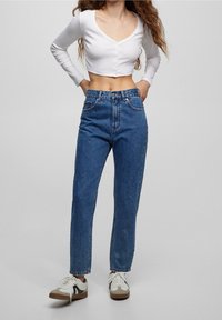 Biały krótki top z długim rękawem i guzikami, zestawiony z niebieskimi dżinsami o wysokim stanie. Sneakersy mają białe i beżowe akcenty.