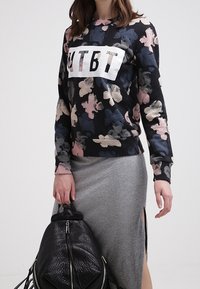 Černá květinová mikina s bílým logem, kombinovaná se šedou sukní s bočním rozparkem. Modelka drží texturovaný černý batoh.