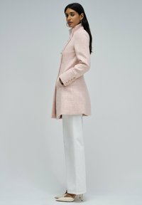 Roze tweed kaput s visokim ovratnikom, zlatnim dugmadi i prilagođenim krojem, uparen s bijelim ravnim hlačama i bež potpeticama.