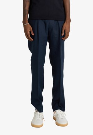 Persona che indossa pantaloni blu navy a gamba dritta, sneakers bianche con suola in gomma e una camicia nera, in piedi davanti a uno sfondo bianco.