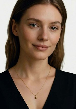 Giovane donna con capelli castani che indossa un top nero e una collana d'oro con un ciondolo a forma di "S", sorridendo dolcemente su uno sfondo semplice.