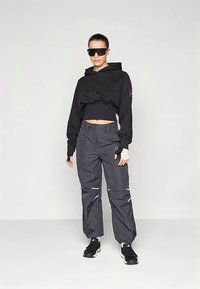 Crna skraćena dukserica s kapuljačom i vrpcama za vezanje, u kombinaciji s tamnosivim hlačama s reflektirajućim prugama i širokim krojem. Crne tenisice upotpunjuju izgled.