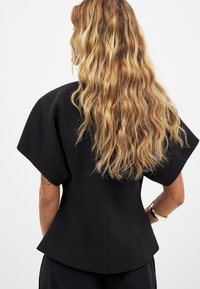 Melnā strukturētā blūze ar īsām, plašām piedurknēm, pieguļošu jostu un gludu tekstūru. Aizskata skats parāda garus, viļņainus matus.