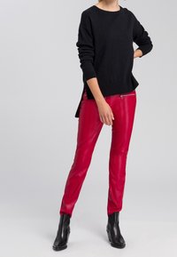 Un pull noir à manches longues associé à un pantalon en cuir rouge ajusté avec des accents de fermeture éclair. Le modèle porte des bottines noires. Fond uni.