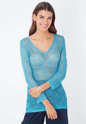 Mujer sonriente con cabello largo y castaño lleva un top de crochet de manga larga turquesa sobre una camiseta interior nude, combinado con pantalones azul marino, de pie frente a un fondo blanco.