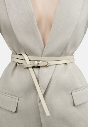 Weißer Ledergürtel mit goldener Schnalle, geschichtetes Design und glatte Textur, getragen über einer beigen Leinenjacke mit tiefem V-Ausschnitt.