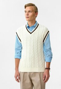 Crème-kleurige gebreide vest met kabelpatronen, V-hals en donkerbruine accenten. Draag het over een lichtblauw gestreept shirt met opgerolde mouwen.