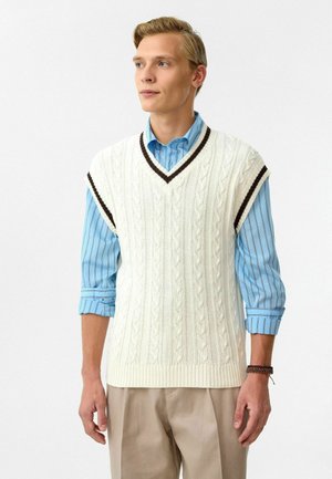 Crème-kleurige gebreide vest met kabelpatronen, V-hals en donkerbruine accenten. Draag het over een lichtblauw gestreept shirt met opgerolde mouwen.
