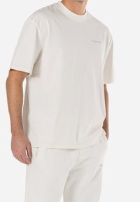 Hombre vestido con una camiseta blanca de manga corta y pantalones blancos a juego, de pie con una mano en el bolsillo frente a un fondo blanco.