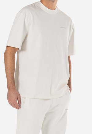 Homme portant un t-shirt blanc à manches courtes et un pantalon blanc assorti, se tenant avec une main dans la poche devant un fond blanc.
