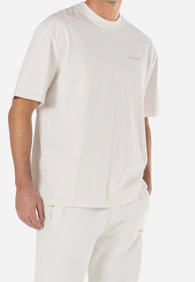 Hombre vestido con una camiseta blanca de manga corta y pantalones blancos a juego, de pie con una mano en el bolsillo frente a un fondo blanco.