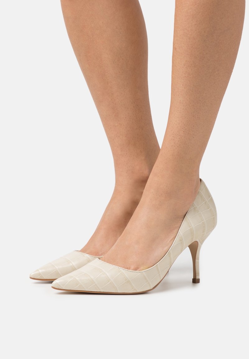 Jambes portant des escarpins beige à bout pointu embossés effet croco, avec des talons fins de 5 cm sur fond blanc.