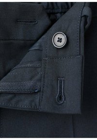 Ciemna tkanina posiada kieszeń na zamek, zapięcie na guzik oraz ozdobne przeszycia. Gładka tekstura z dopasowanym krojem.