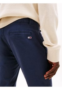 Primer plano de una persona que lleva pantalones azul marino con un pequeño logo en el bolsillo trasero y un suéter de punto color crema, con la mano descansando a un lado.