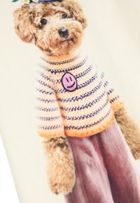 Un chien portant un pull à rayures avec des bordures orange et un patch de smiley rose, texture duveteuse, se tenant devant un fond clair.