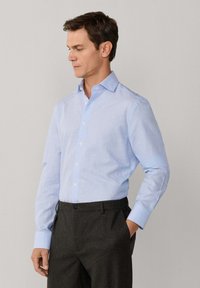 Homme portant une chemise bleu clair à boutons et un pantalon gris foncé, debout avec une main dans la poche devant un fond uni.