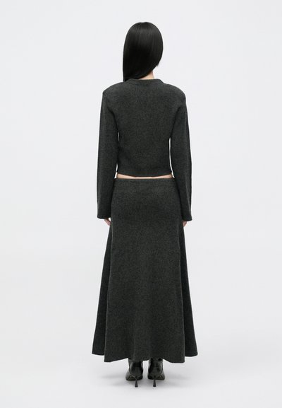 Γυναίκα με μακριά μαύρα μαλλιά, φορώντας ένα σκούρο γκρι κοντό πουλόβερ με μακριά μανίκια, ταιριαστό maxi φούστα και μαύρες ψηλές μπότες αστραγάλου, γυρισμένη προς το μέρος.