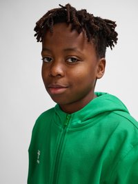 Garçon avec de courtes dreadlocks portant un sweat à capuche vert zippé, regardant légèrement sur le côté sur un fond clair uni.