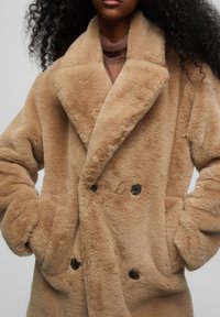 Abrigo beige de doble botonadura hecho de un tejido suave y esponjoso. Presenta un collar con muesca, botones grandes y bolsillos laterales con un acabado texturizado.