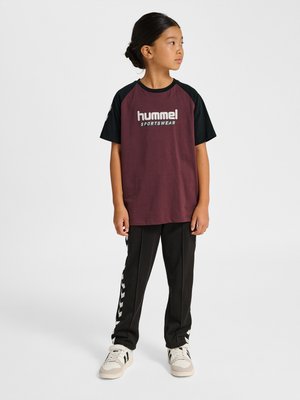Child standing, wearing maroon and black hummel sportswear t-shirt, black pants with white chevron stripes, and white sneakers, looking right.