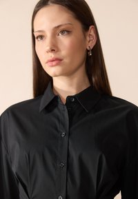 Camicia nera con bottoni realizzata in tessuto liscio, caratterizzata da un collo classico, una leggera lucentezza e sei bottoni frontali.